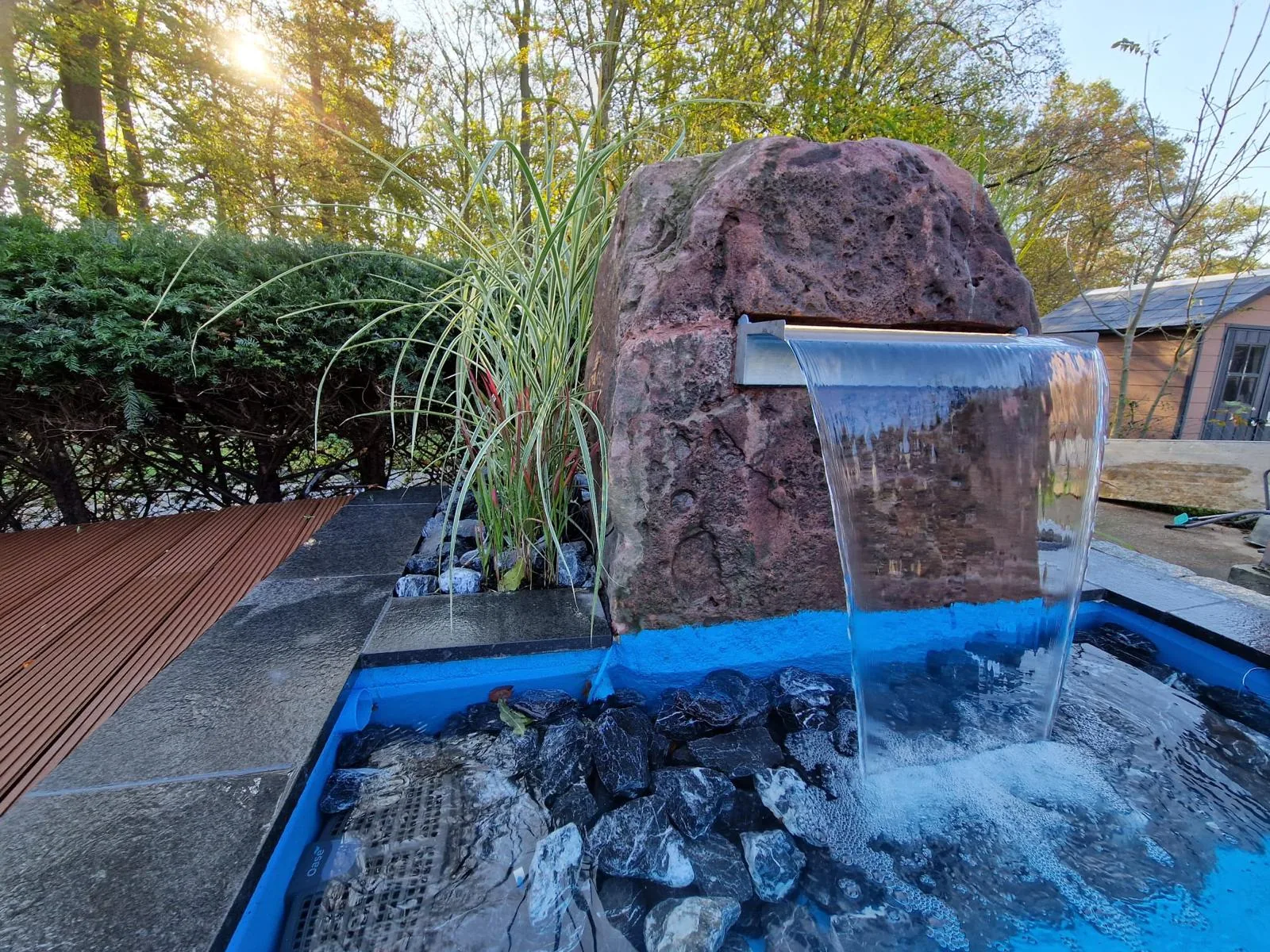 Wasserspiel aus rötlichem Naturstein mit Edelstahl-Wasserfall in einen Teich